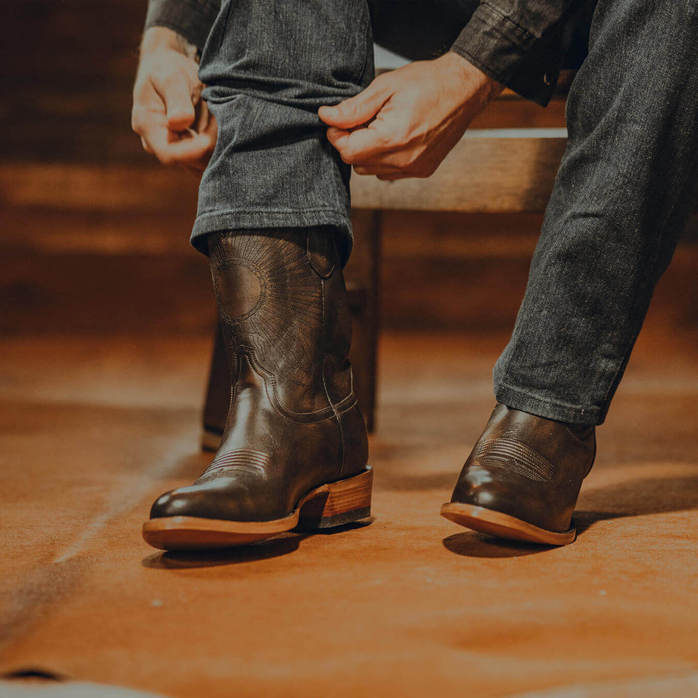 Person adjusting jeans over well-fitted cowboy boots—dark leather with stitching detail, showing snug fit around the instep and shaft on a rustic floor.
